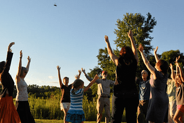 A group dances at the edge of woods and prairie, in a circle, hands raising to the sky where a bird if flying by.