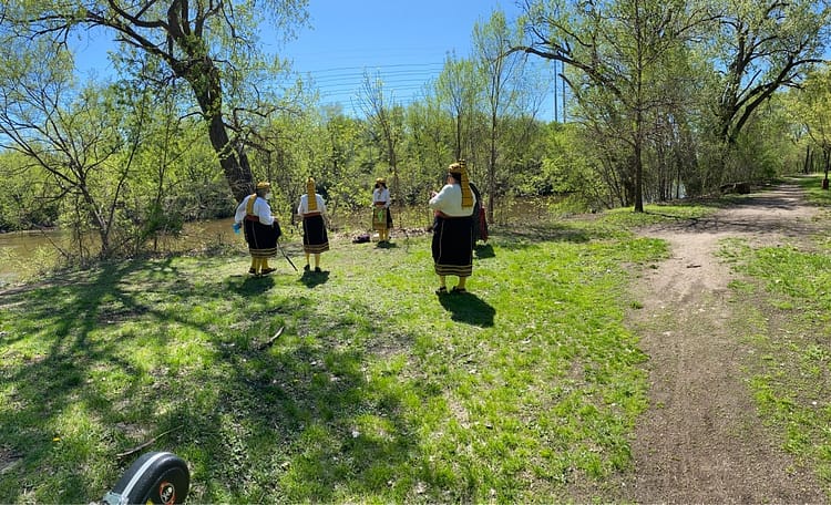 Dancers gather on spring green grass by a murky river.