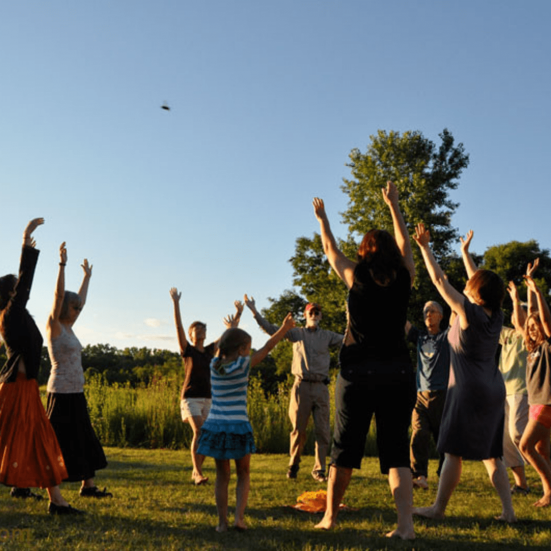 A group dances at the edge of woods and prairie, in a circle, hands raising to the sky where a bird if flying by. 